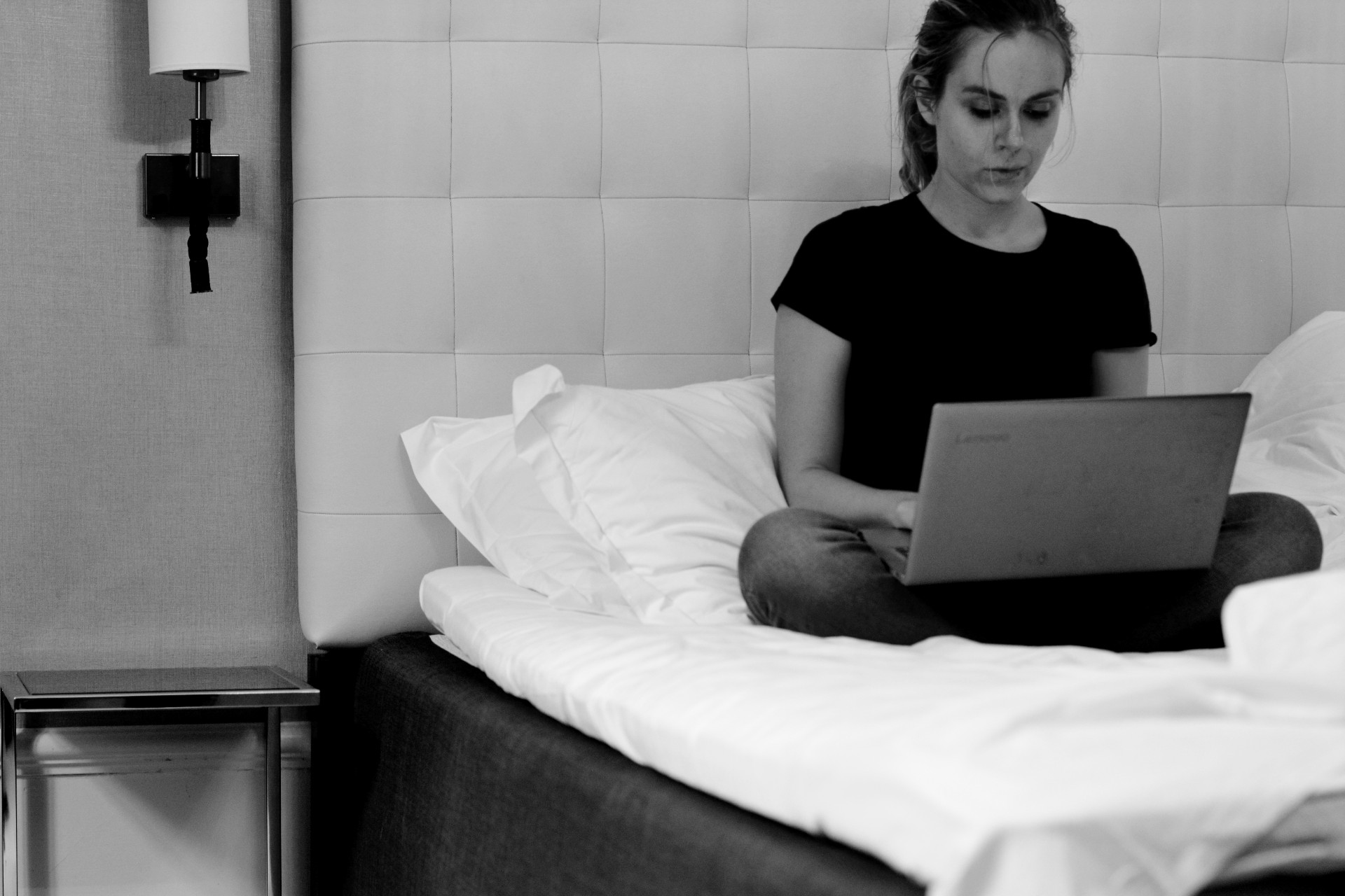 Ulrika skriver på en laptop på en hotellsäng. Svartvit bild.