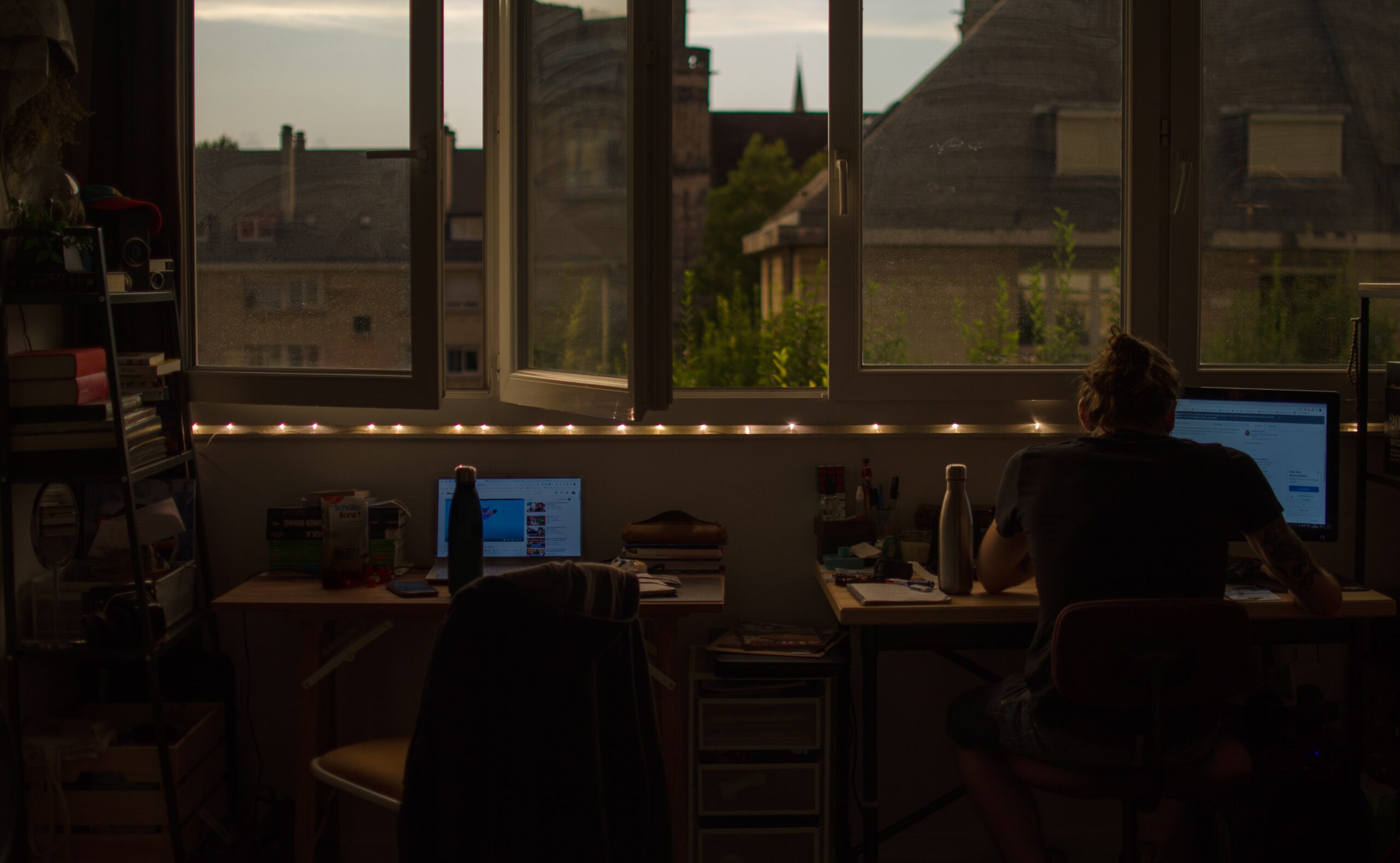 Två skrivbord framför öppet fönster med ljusslingor i fönstret och fin utsikt över hustak. En man sitter vid ett skrivbord och jobbar.