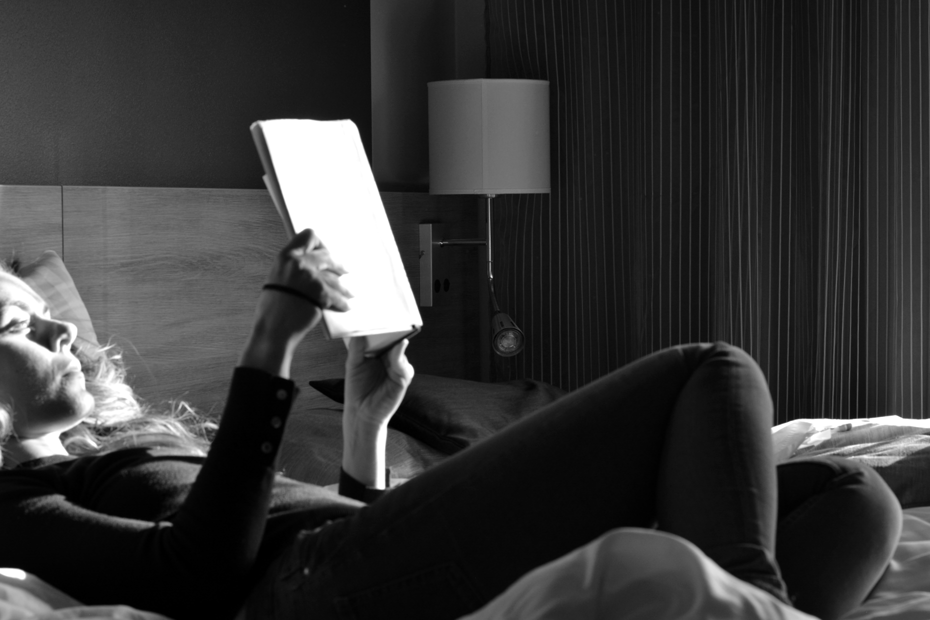 Ulrika Nettelblad ligger i hotellsäng på Hotell Pommern och läser bok. Svartvit bild, motljus, ansiktet kraftig upplyst av solen.