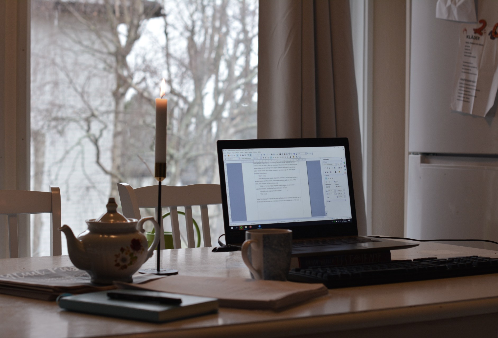 Laptop, tekanna, romanmanus utskrivet på papper på köksbord framför fönster.
