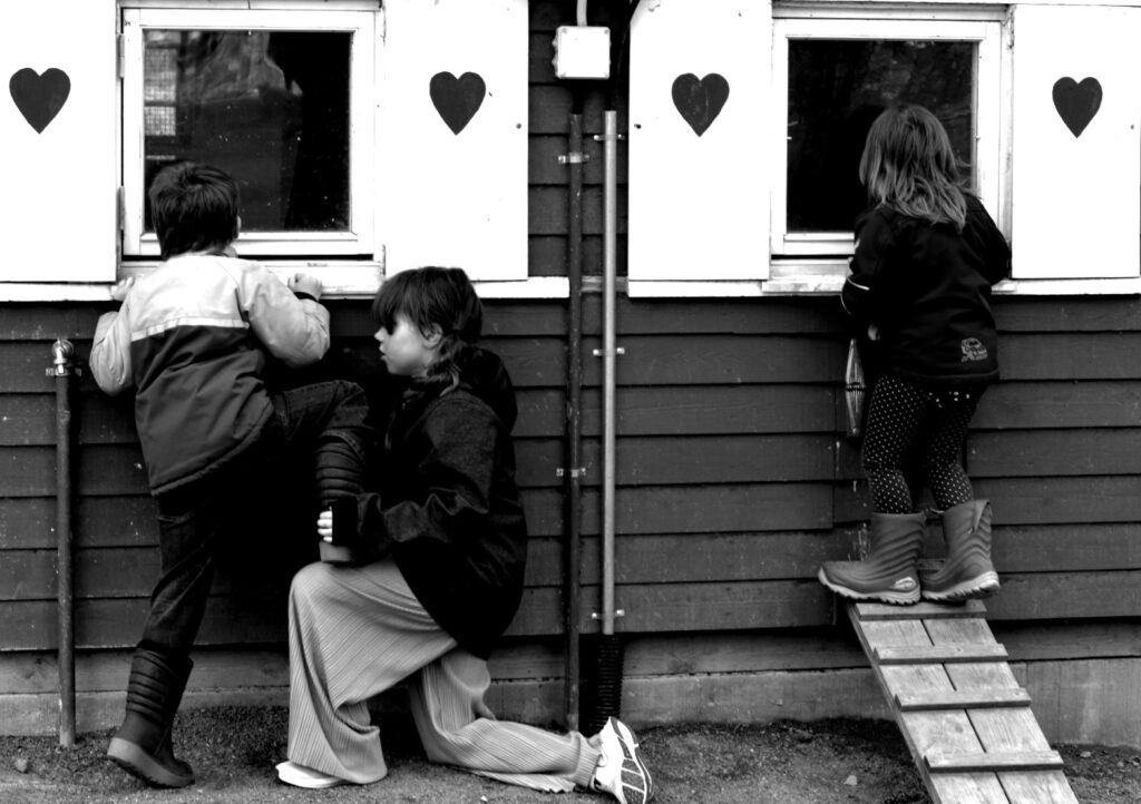 Tre barn framför fönster med hjärtan på. Ett barn får stå på ett annat barns knä för att kunna titta in i ett fönster. Svartvit bild.